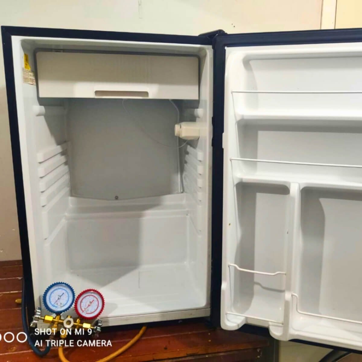 Frigobar branco aberto com manômetro manifold conectado para recarga de gás e teste de pressão.